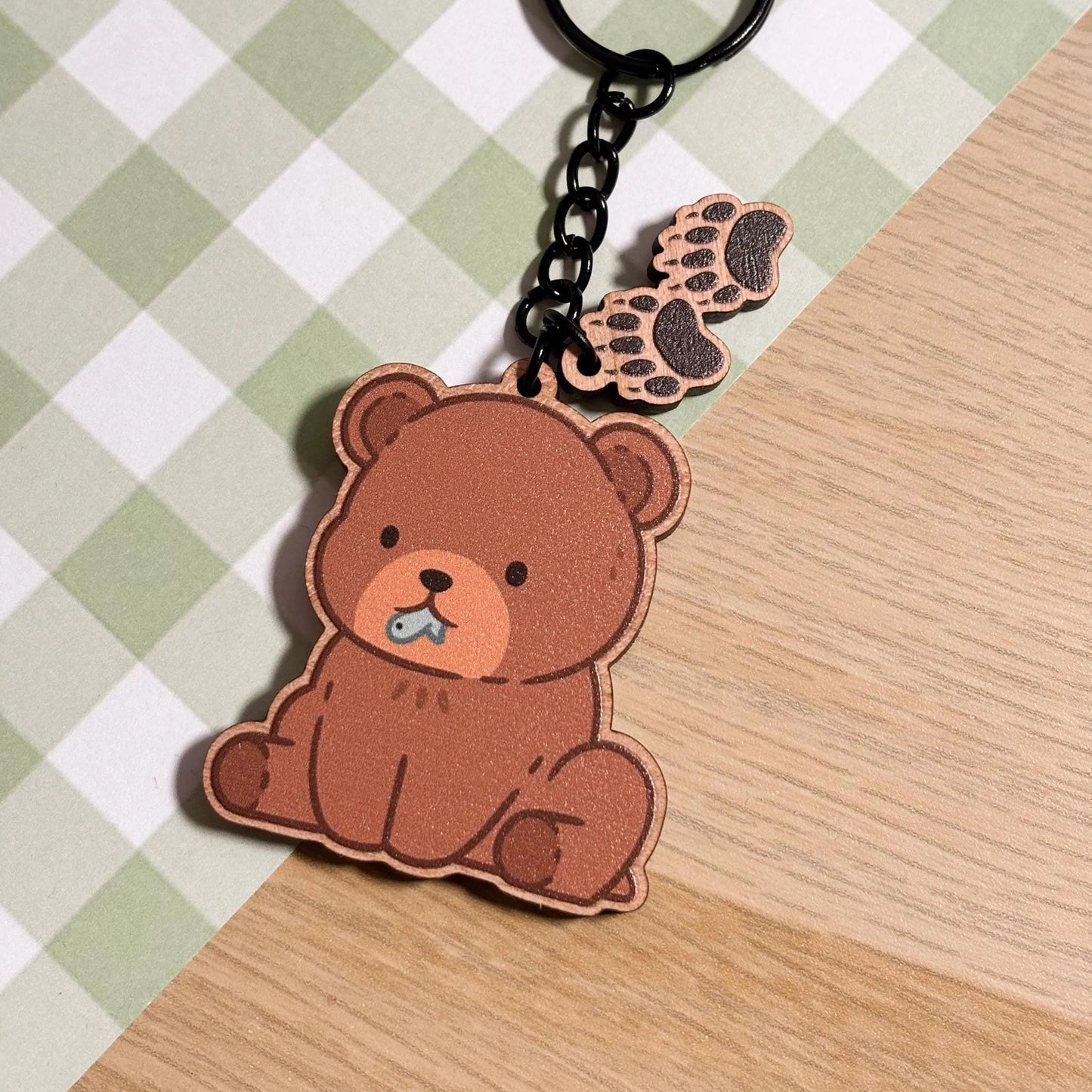 A charming wooden keychain on a green and white checkered surface. The main charm is a cute, seated brown bear with a light brown muzzle and a tiny blue fish in its mouth. Two smaller wooden charms shaped like bear paw prints dangle from the keychain's black chain. The overall aesthetic is sweet and whimsical.