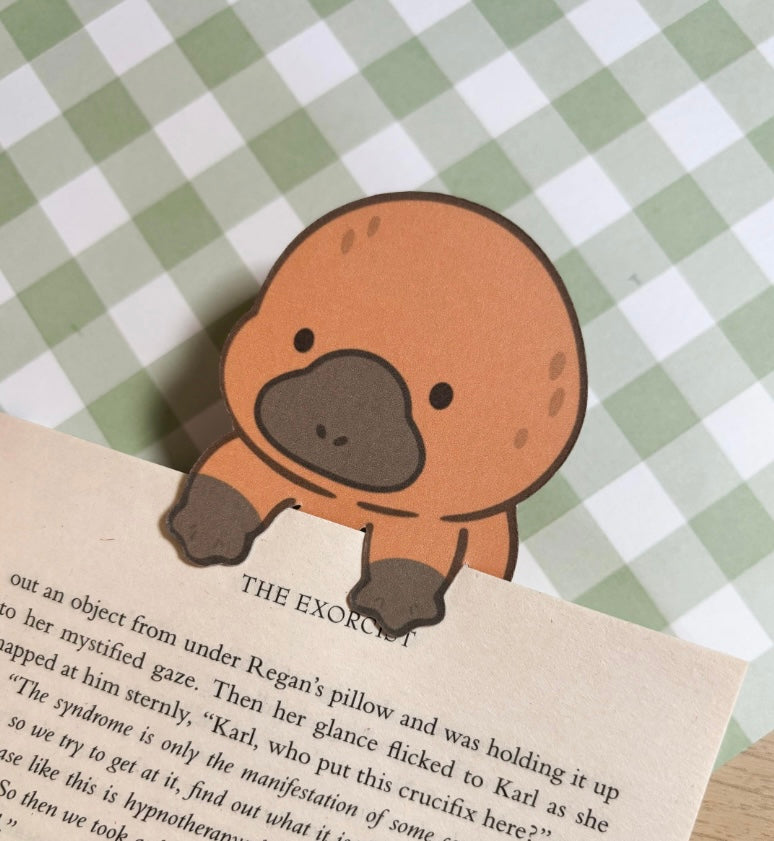 A brown platypus bookmark with a black nose and eyes. The bookmark is holding a book open to a page