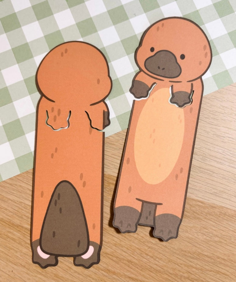 Two brown platypus bookmark cutouts. The platypus on the left is facing away from the viewer and the platypus on the right is facing the viewer. Both platypus have a brown body, a black nose, and white feet.