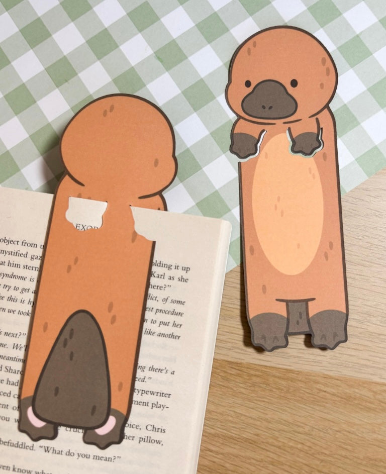 Two brown and tan platypus bookmark cutouts. The platypus are standing on a book. The platypus are facing forward, The platypus are holding their front paws in front of them.