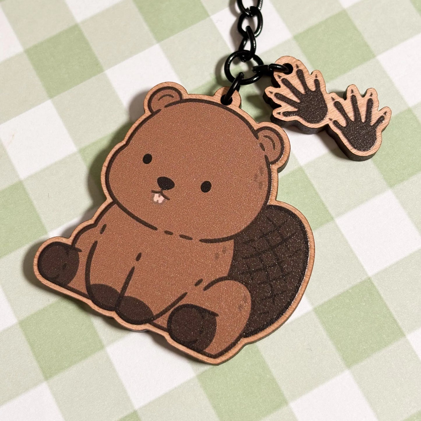 A delightful wooden keychain featuring a cartoon beaver rests on a light green and white checkered background. The seated beaver charm has a textured, paddle-shaped tail and two small, visible front teeth. Two smaller wooden charms shaped like webbed beaver paws dangle from the black metal chain and ring of the keychain. The overall impression is whimsical and endearing.