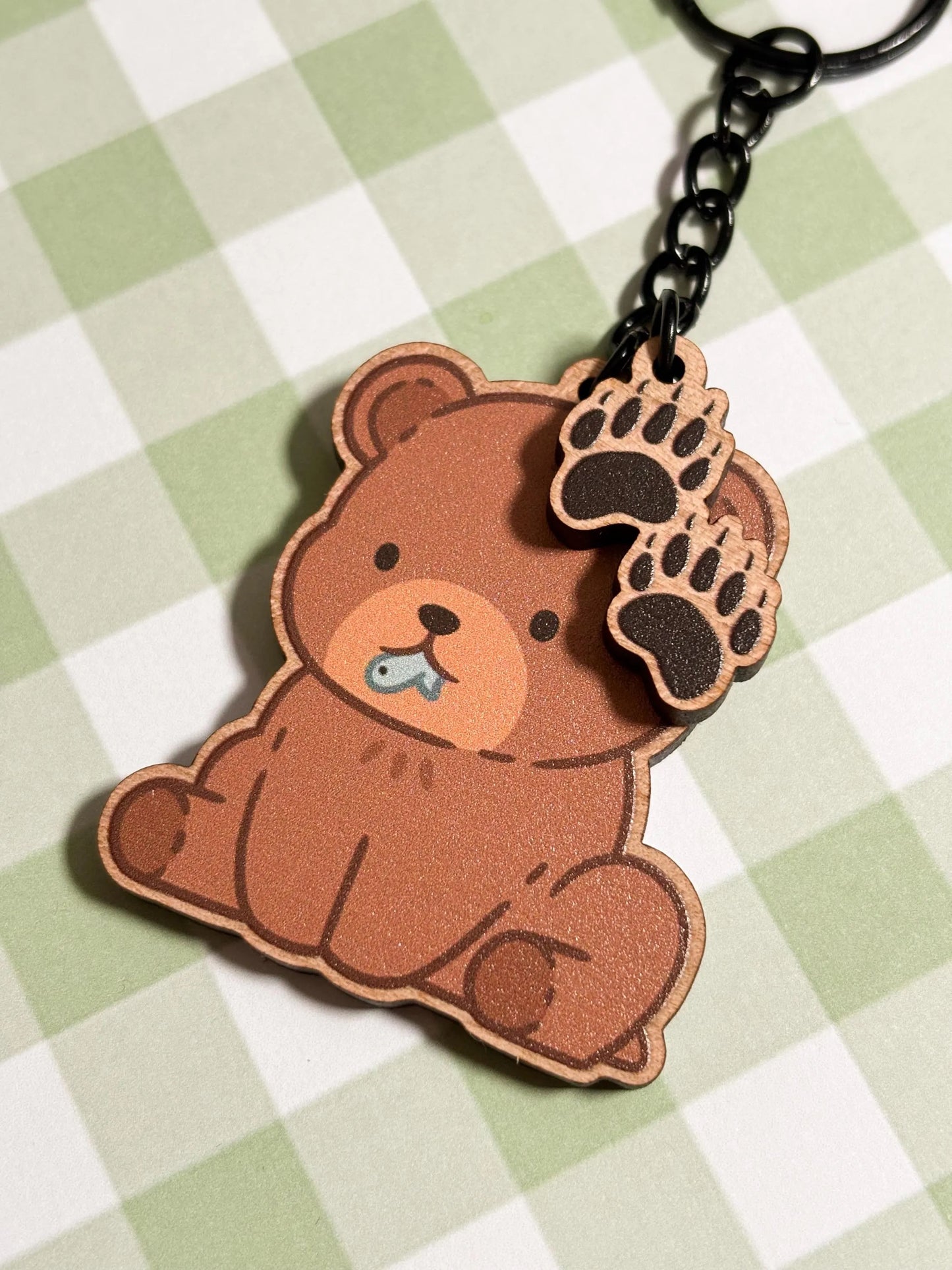 a cute brown bear charm sits with a tiny blue fish in its mouth, accompanied by two smaller paw print charms dangling above. close up.