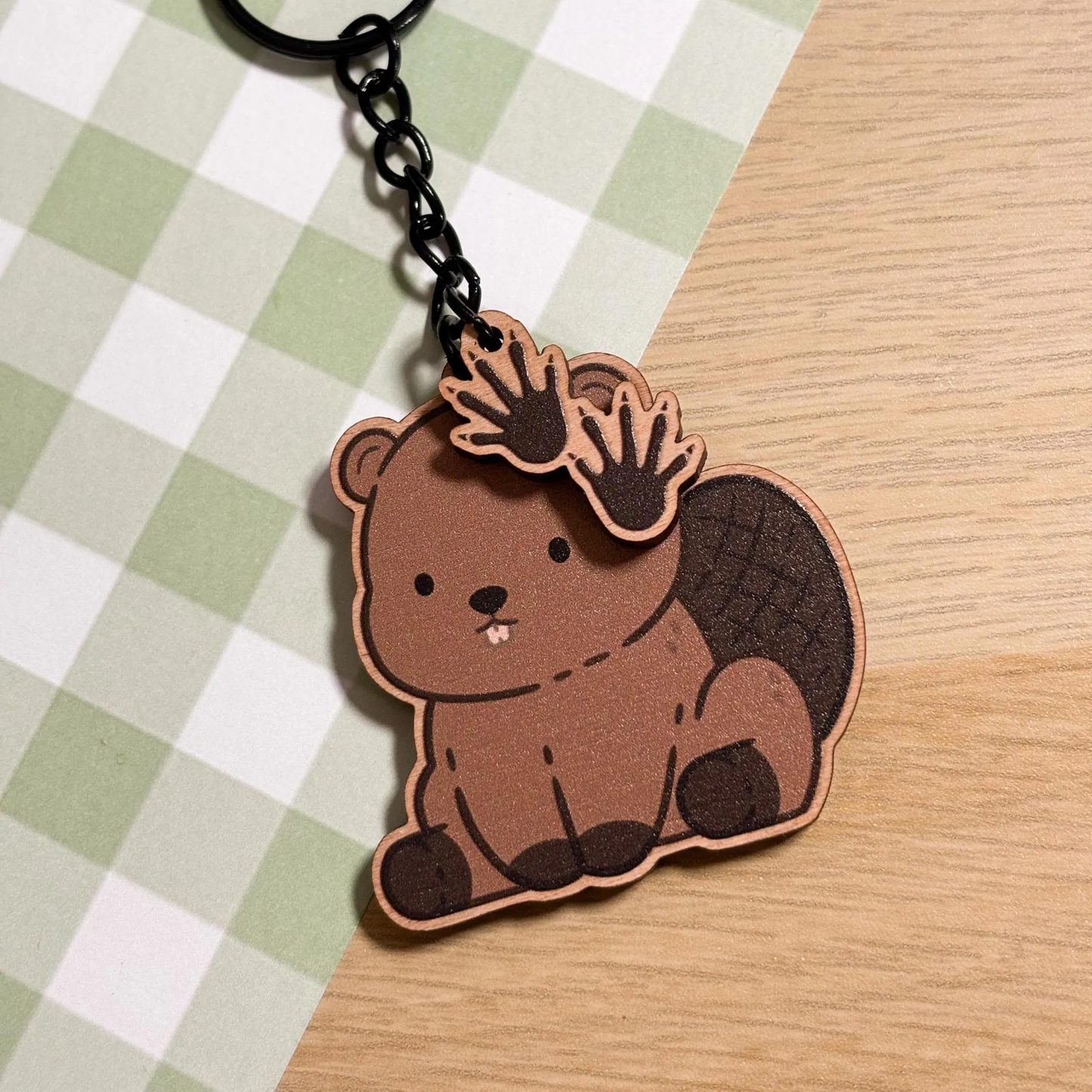 A charming wooden keychain of a cartoon beaver is displayed on a light green and white checkered surface. The beaver is depicted in a seated position, showcasing its flat, textured tail and two prominent front teeth. Two smaller wooden charms shaped like webbed beaver paws dangle from the black metal chain attached to the main beaver charm. The style is cute and illustrative.