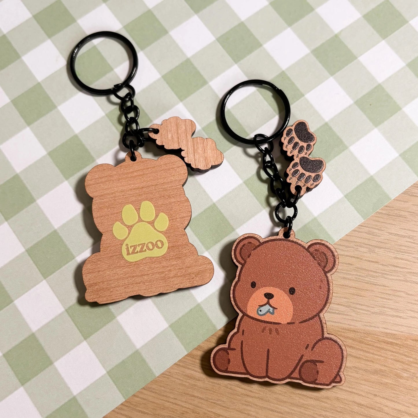 Two adorable wooden keychains rest on a light green and white checkered background. On the right, a brown bear charm sits with a tiny blue fish in its mouth, accompanied by two smaller paw print charms dangling above. To the left, another bear-shaped charm features a muted light green paw print and the word "izzoo" in playful lettering. Both keychains have black metal rings and chains. The scene is bright and cheerful.