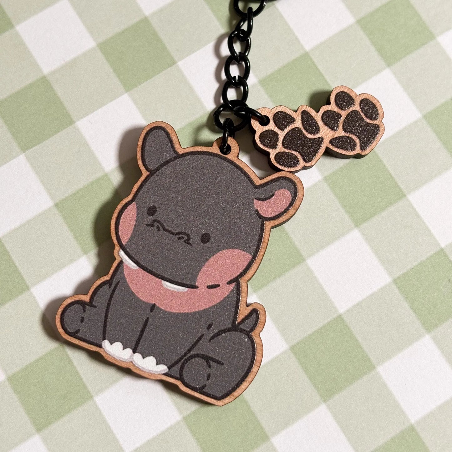 A charming wooden keychain of a cartoon hippopotamus is pictured on a light green and white checkered surface. The hippo is depicted in a seated pose, featuring a grey body, rosy pink cheeks and inner ears, and two small white teeth peeking out. A smaller wooden charm shaped like a paw print with four rounded toes dangles from the black metal chain and ring of the keychain. The style is cute and playful.