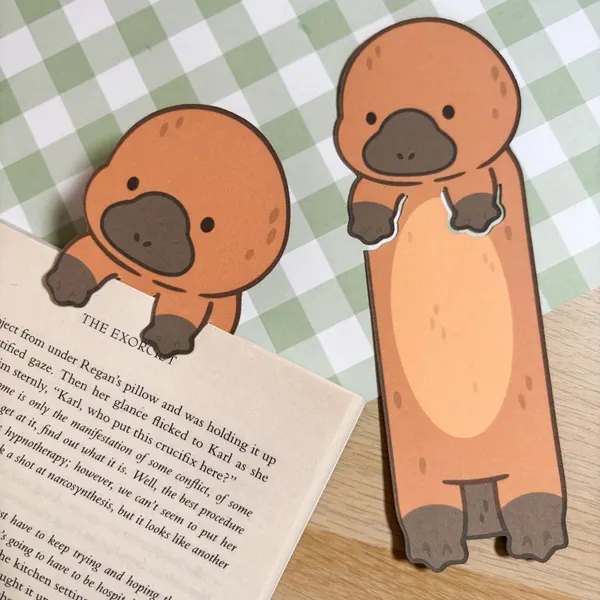 Two brown platypus bookmark designs. The platypus the left is holding a book. The platypus on the right is standing upright. Both platypus have a brown body with a black nose and feet.