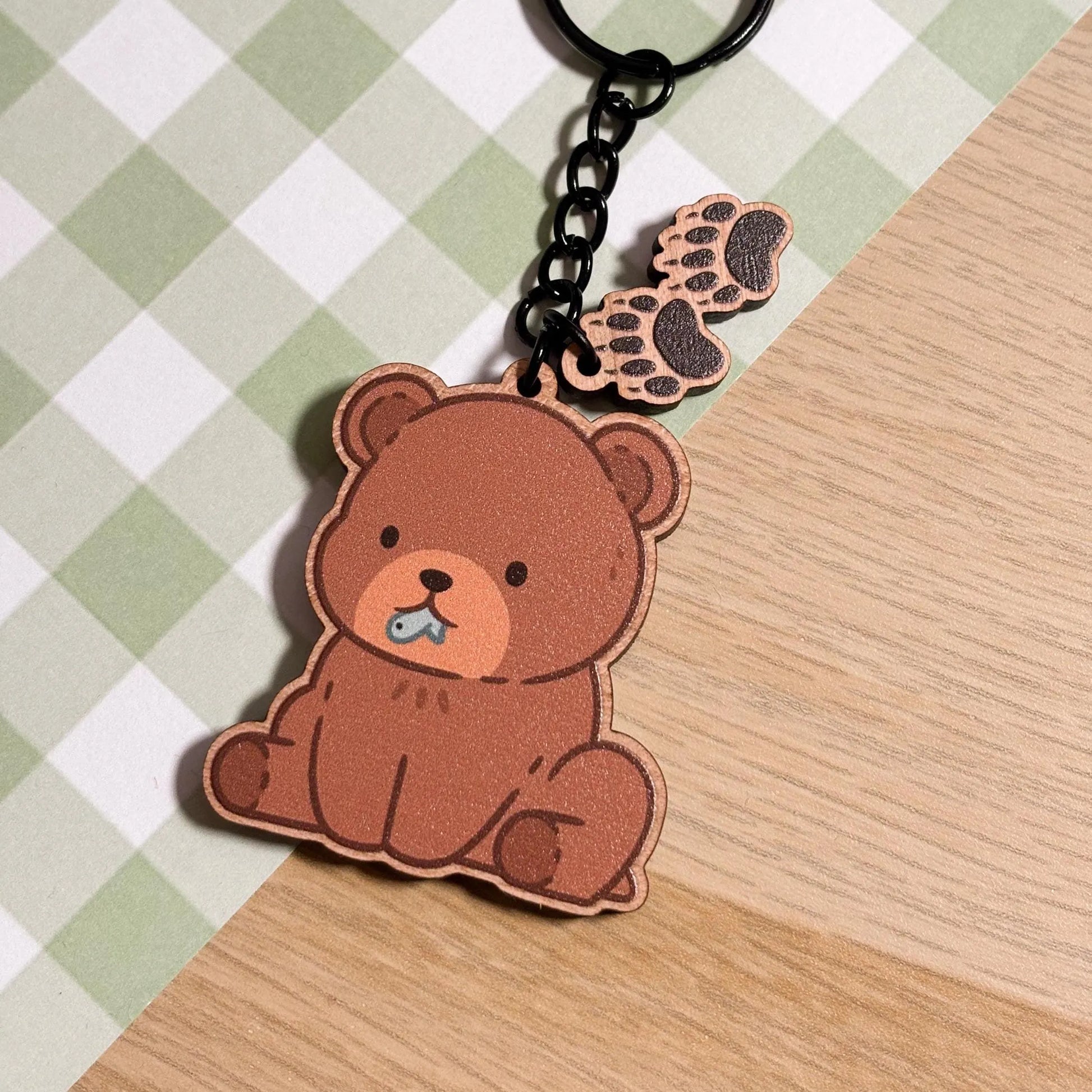 A charming wooden keychain on a green and white checkered surface. The main charm is a cute, seated brown bear with a light brown muzzle and a tiny blue fish in its mouth. Two smaller wooden charms shaped like bear paw prints dangle from the keychain's black chain. The overall aesthetic is sweet and whimsical.