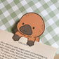 A brown platypus bookmark with a black nose and eyes. The bookmark is holding a book open to a page