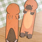 Two brown platypus bookmark cutouts. The platypus on the left is facing away from the viewer and the platypus on the right is facing the viewer. Both platypus have a brown body, a black nose, and white feet.