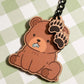a cute brown bear charm sits with a tiny blue fish in its mouth, accompanied by two smaller paw print charms dangling above. close up.