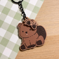 A charming wooden keychain of a cartoon beaver is displayed on a light green and white checkered surface. The beaver is depicted in a seated position, showcasing its flat, textured tail and two prominent front teeth. Two smaller wooden charms shaped like webbed beaver paws dangle from the black metal chain attached to the main beaver charm. The style is cute and illustrative.