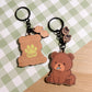 Two adorable wooden keychains rest on a light green and white checkered background. On the right, a brown bear charm sits with a tiny blue fish in its mouth, accompanied by two smaller paw print charms dangling above. To the left, another bear-shaped charm features a muted light green paw print and the word "izzoo" in playful lettering. Both keychains have black metal rings and chains. The scene is bright and cheerful.