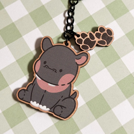 A charming wooden keychain of a cartoon hippopotamus is pictured on a light green and white checkered surface. The hippo is depicted in a seated pose, featuring a grey body, rosy pink cheeks and inner ears, and two small white teeth peeking out. A smaller wooden charm shaped like a paw print with four rounded toes dangles from the black metal chain and ring of the keychain. The style is cute and playful.