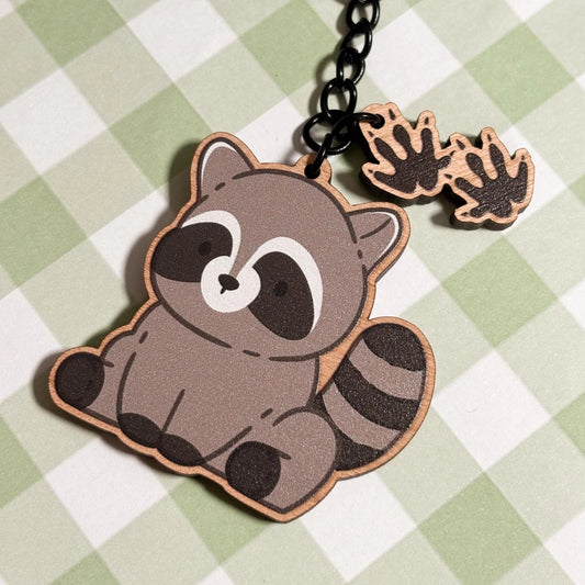 A delightful wooden keychain of a cartoon raccoon sits on a light green and white checkered surface. The seated raccoon has its characteristic black and white striped tail and a black mask around its eyes. Two smaller wooden charms shaped like raccoon paw prints with five distinct toes dangle from the black metal chain and ring of the keychain. The overall style is endearing and whimsical.