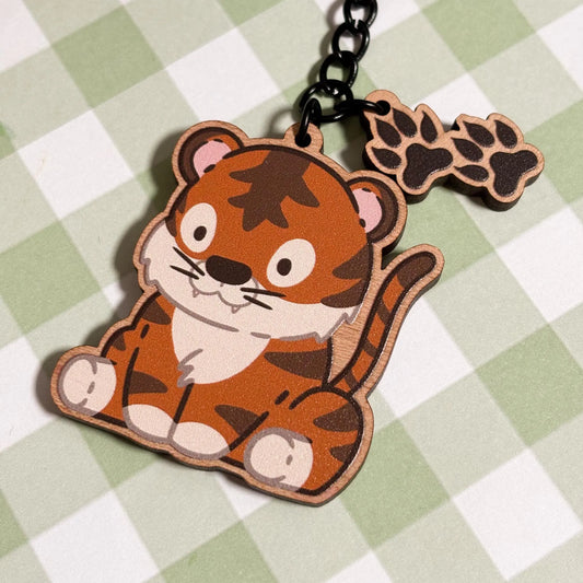 A vibrant wooden keychain of a cartoon tiger is displayed on a light green and white checkered surface. The seated tiger has bright orange fur with dark brown stripes, a white belly and muzzle, and small white teeth showing. Two smaller wooden charms shaped like tiger paw prints with four rounded toes dangle from the black metal chain and ring of the keychain. The style is playful and full of character.